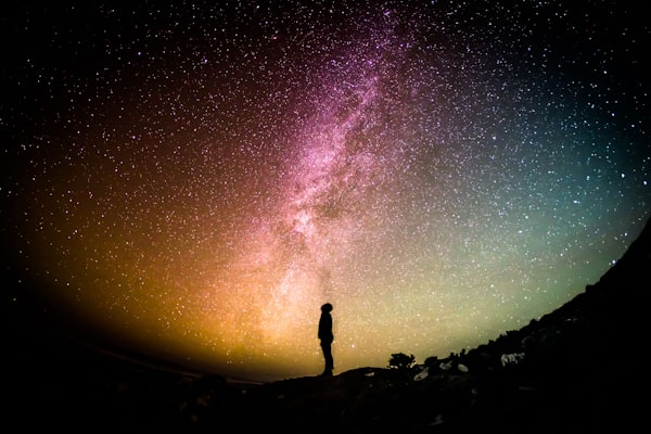A lone human figure silhouetted against the Milky Way and a star-filled night sky.